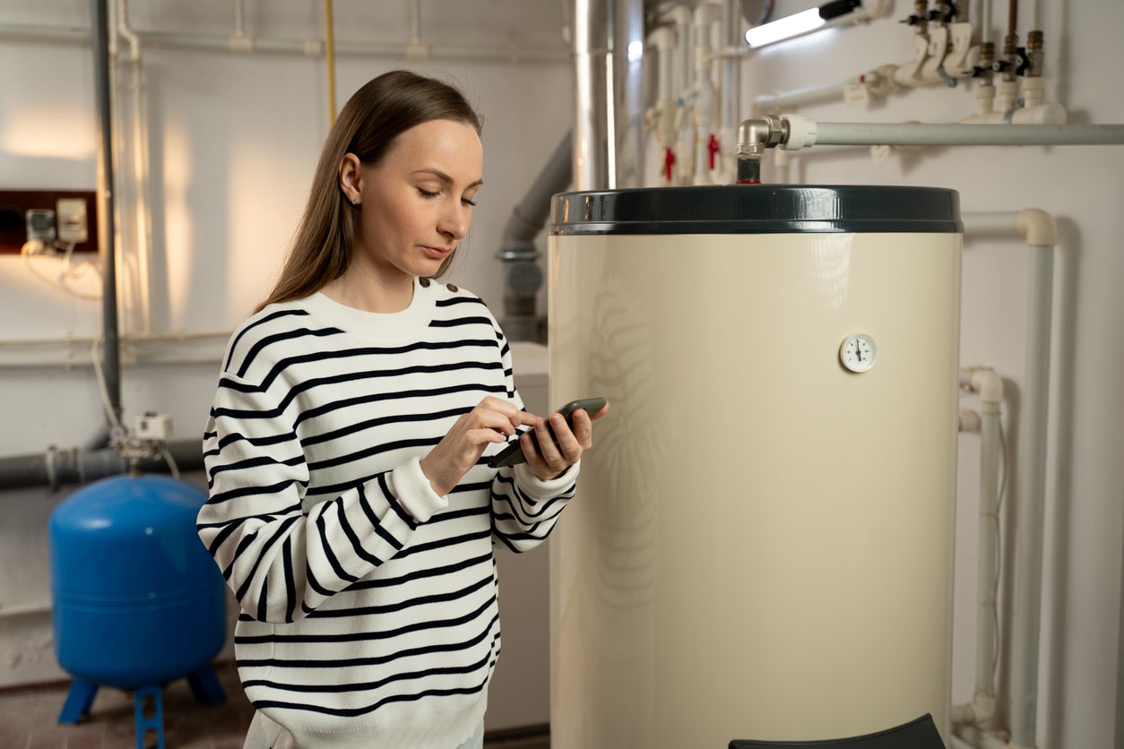 A female homeowner is examining her home’s water heater while holding her phone to call for repairs.