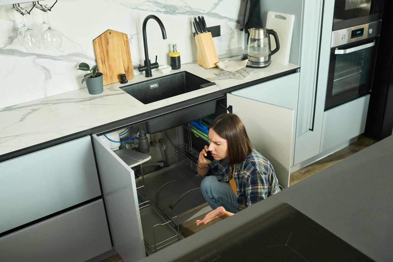 A homeowner calls a residential plumber while inspecting the pipes under her leaking kitchen sink.